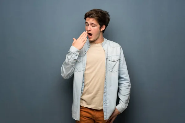 Teenager man with jean jacket over grey wall yawning and covering wide open mouth with hand