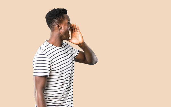 Dark skinned man with striped shirt shouting with mouth wide open and announcing something on isolated ocher background