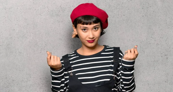 Young woman with beret making money gesture over textured wall