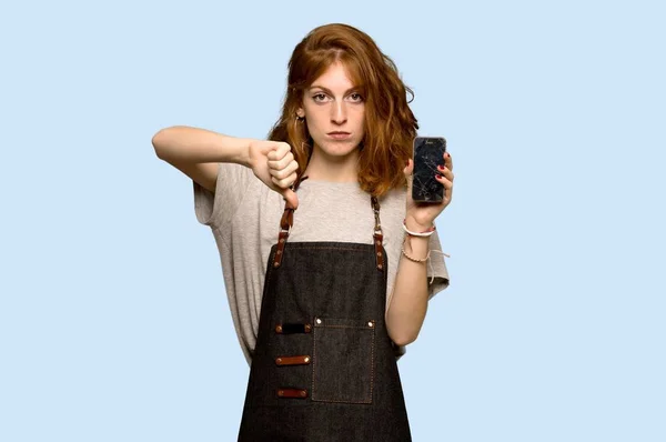 青い背景に壊れた携帯電話をエプロンの若い赤毛の女性 — ストック写真