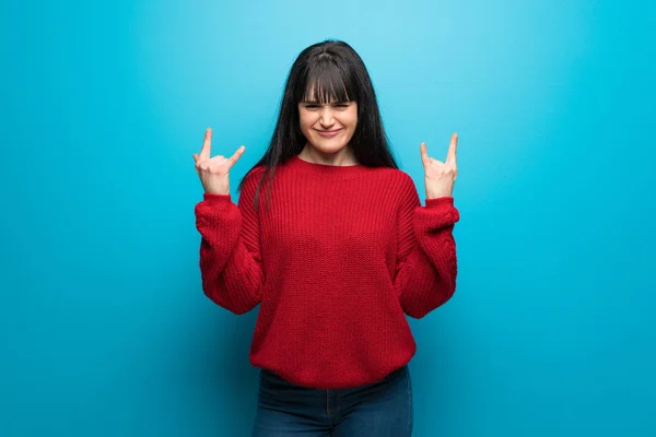 Woman with red sweater over blue wall making rock gesture