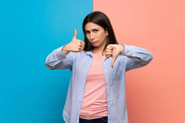 Young woman over pink and blue wall making good-bad sign. Undecided between yes or not