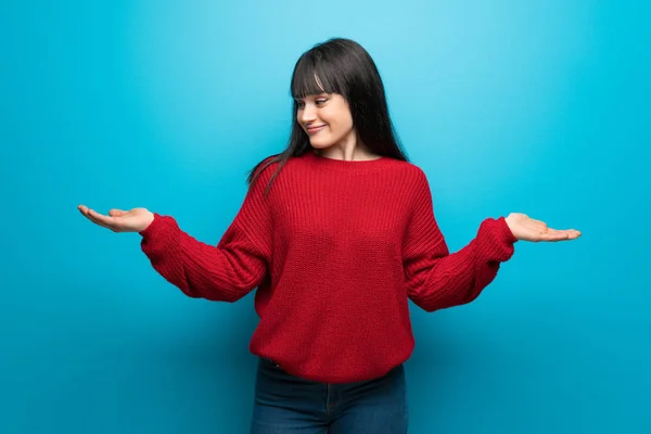 Woman with red sweater over blue wall holding copyspace with two hands