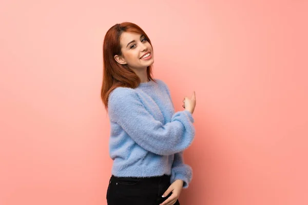 Young redhead woman over pink background pointing back with the index finger
