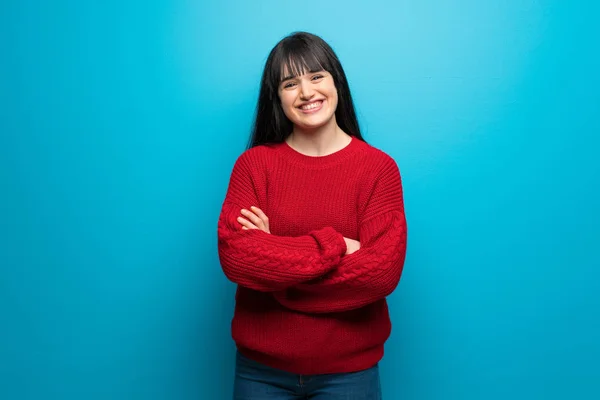Woman with red sweater over blue wall keeping the arms crossed in frontal position