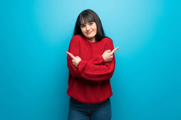 Woman with red sweater over blue wall pointing to the laterals having doubts