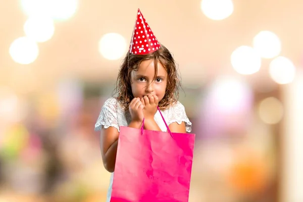 La bambina a una festa di compleanno con in mano una busta regalo è un po 'nervosa e spaventata su uno sfondo sfocato. — Foto Stock