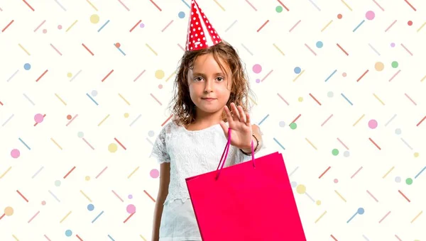 Menina em uma festa de aniversário segurando um saco de presente fazendo parar gesto com a mão no fundo colorido — Fotografia de Stock