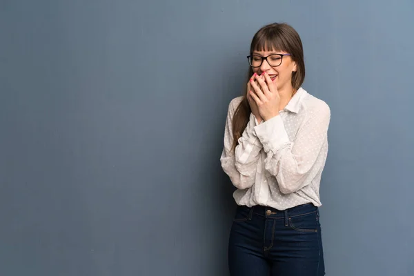 Woman with glasses over blue wall smiling a lot while covering mouth