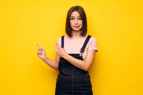 Young woman over yellow wall frightened and pointing to the side
