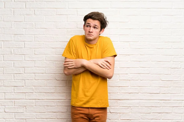 Teenager man over white brick wall making doubts gesture while lifting the shoulders