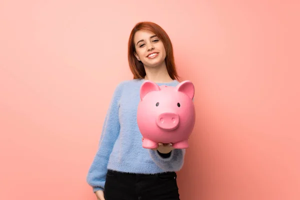 Young redhead woman over pink background holding a piggybank