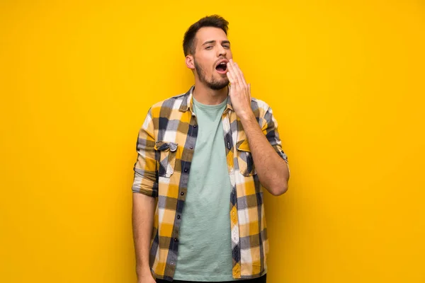 Handsome man over yellow wall yawning and covering wide open mouth with hand