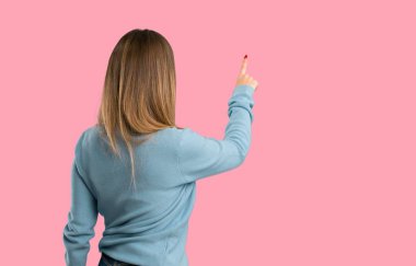 Mavi gömlek geri izole pembe bir arka plan üzerinde işaret parmağı ile işaret ile sarışın kadın