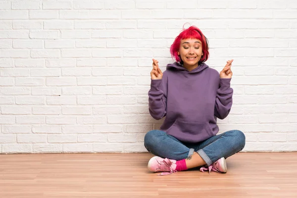 Young woman with pink hair sitting on the floor with fingers crossing and wishing the best