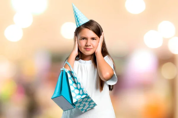 Menina em uma festa de aniversário segurando um saco de presente cobrindo ambas as orelhas com as mãos em fundo desfocado — Fotografia de Stock