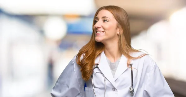 红发医生妇女摆姿势与手臂在臀部和笑在医院 — 图库照片
