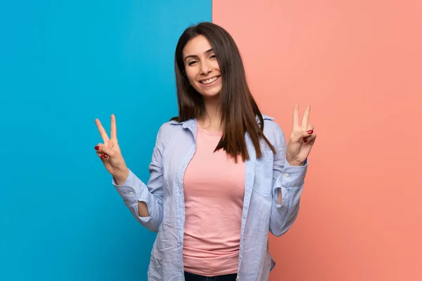 Young woman over pink and blue wall smiling and showing victory sign with both hands