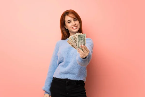 Young redhead woman over pink background taking a lot of money