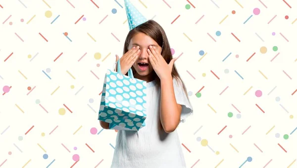 Menina em uma festa de aniversário segurando um saco de presente cobrindo os olhos por mãos em fundo colorido — Fotografia de Stock