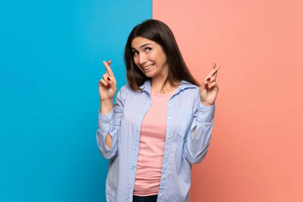 Young woman over pink and blue wall with fingers crossing and wishing the best