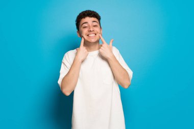 Mutlu ve keyifli bir ifadeyle gülümseyen mavi arka plan üzerinde genç adam