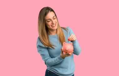 Mavi gömlek izole pembe arka plan dolu olduğu için kumbara ve mutlu çekici sarışın kadınla