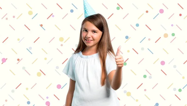 Menina em uma festa de aniversário segurando um saco de presente dando um gesto polegares para cima e sorrindo no fundo colorido — Fotografia de Stock