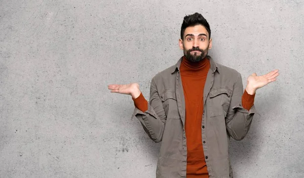 Handsome man with beard having doubts while raising hands and shoulders over textured wall