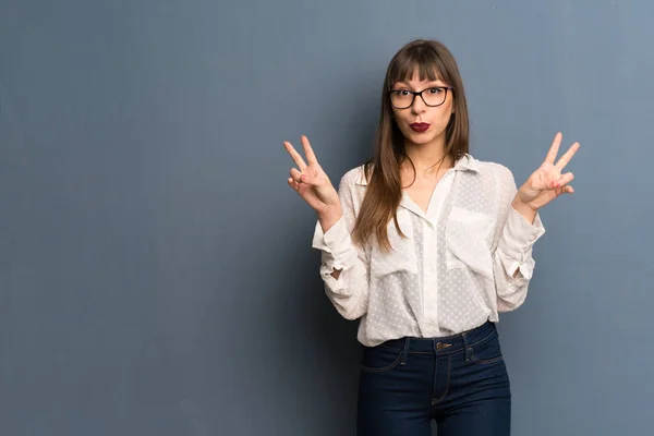 Woman with glasses over blue wall smiling and showing victory sign with both hands