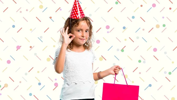 Bambina ad una festa di compleanno con una borsa regalo che mostra un segno ok con le dita su sfondo colorato — Foto Stock