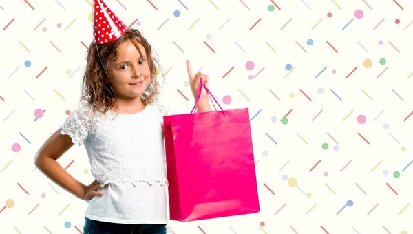 Menina em uma festa de aniversário segurando um saco de presente contando número um sinal no fundo colorido — Fotografia de Stock