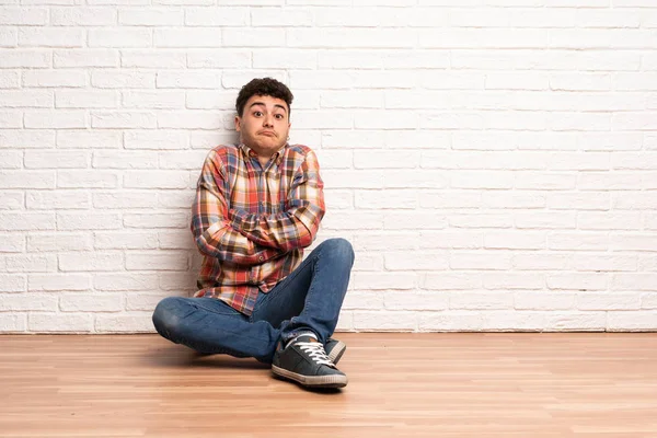 Mladý Muž Sedící Podlaze Dělá Pochybovačné Gesto Zatímco Zvedá Ramena — Stock fotografie