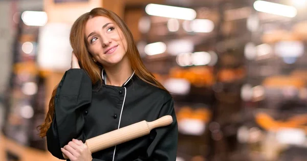 Young redhead chef woman thinking an idea while scratching head in the bakery