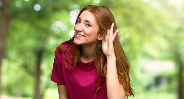 年轻的红头发的女孩在公园里把手放在耳朵上听东西 — 图库照片