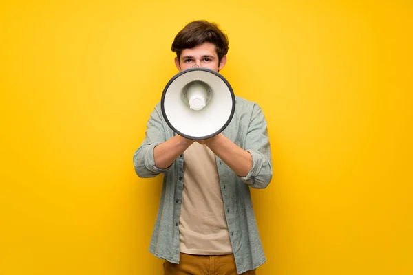 Teenager man over yellow wall shouting through a megaphone to announce something