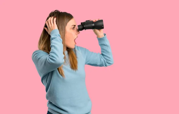 Blonde woman with blue shirt and looking in the distance with binoculars on isolated pink background