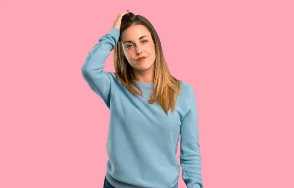 Blonde woman with blue shirt with an expression of frustration and not understanding on isolated pink background