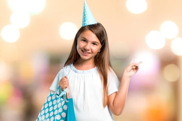 Niña en una fiesta de cumpleaños sosteniendo una bolsa de regalo sonriendo y mostrando el signo de victoria en un fondo desenfocado —  Fotos de Stock