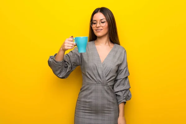 Young woman with glasses over yellow wall holding a hot cup of coffee