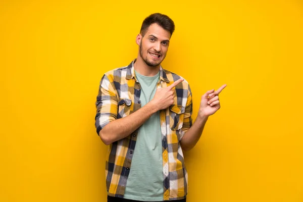 Handsome man over yellow wall frightened and pointing to the side