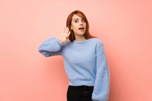 Young redhead woman over pink background listening to something by putting hand on the ear