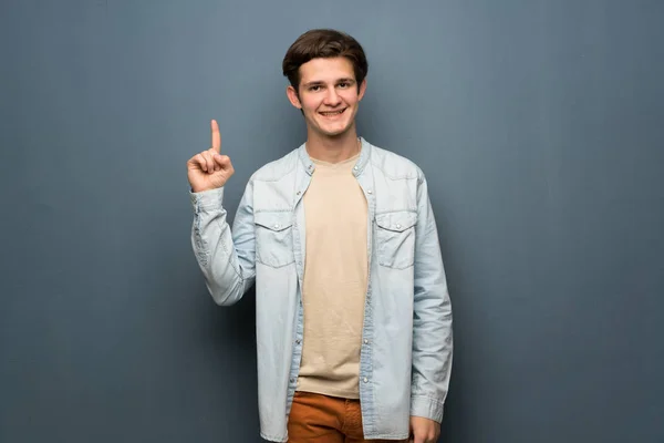 Teenager man with jean jacket over grey wall showing and lifting a finger in sign of the best