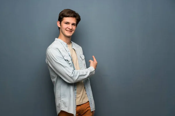 Teenager man with jean jacket over grey wall pointing back