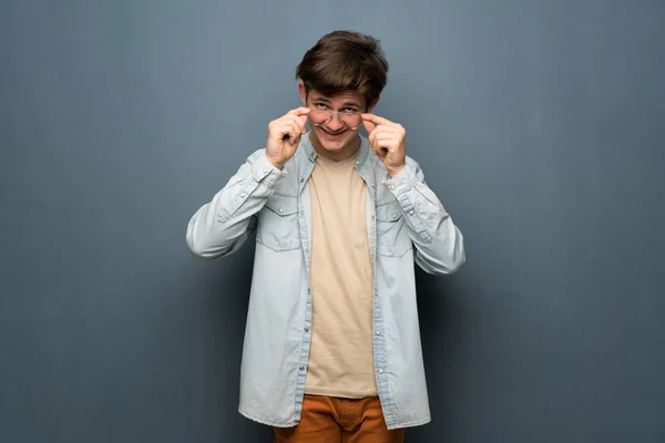 Teenager man with jean jacket over grey wall with glasses and surprised