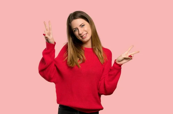 笑みを浮かべて ピンクの背景以上の両方の手で勝利のサインを示す赤いセーターを持つ若い女性 — ストック写真