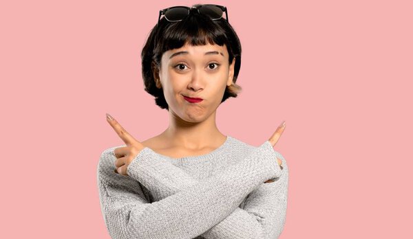 Молодая женщина с короткими волосами, указывающими на боковые стороны, сомневающаяся на изолированном розовом фоне
