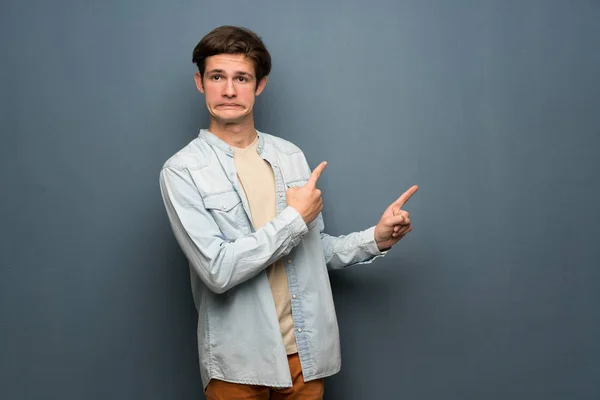 Teenager man with jean jacket over grey wall frightened and pointing to the side