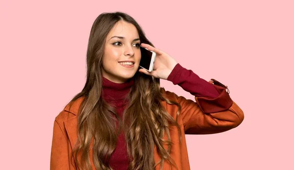 Young woman with coat keeping a conversation with the mobile phone over isolated pink background