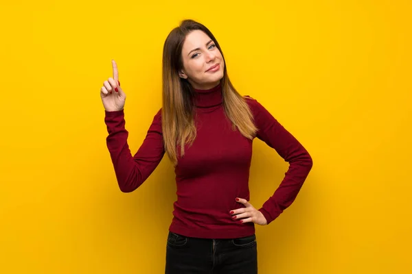 妇女与高领毛衣在黄色的墙壁显示和举起一个手指在最好的标志 — 图库照片